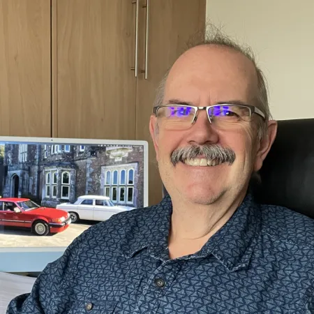 Someone smiling in purple glasses and blue shirt, sitting at desk with city view in background