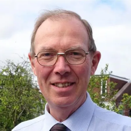 Professional headshot of person in glasses and blue dress shirt with tie against outdoor background