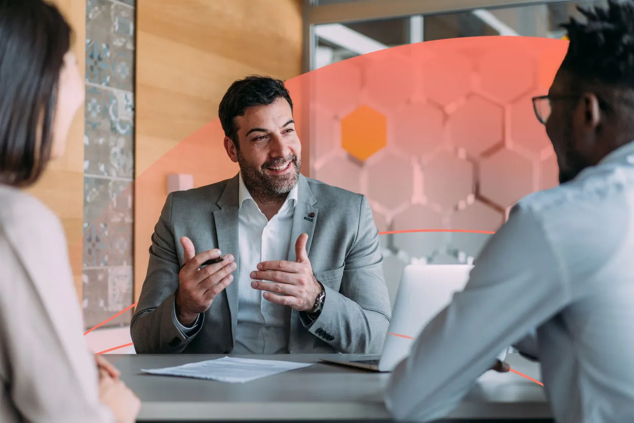 A man in a suit speaking to two colleagues across a table, leading a business discussion.