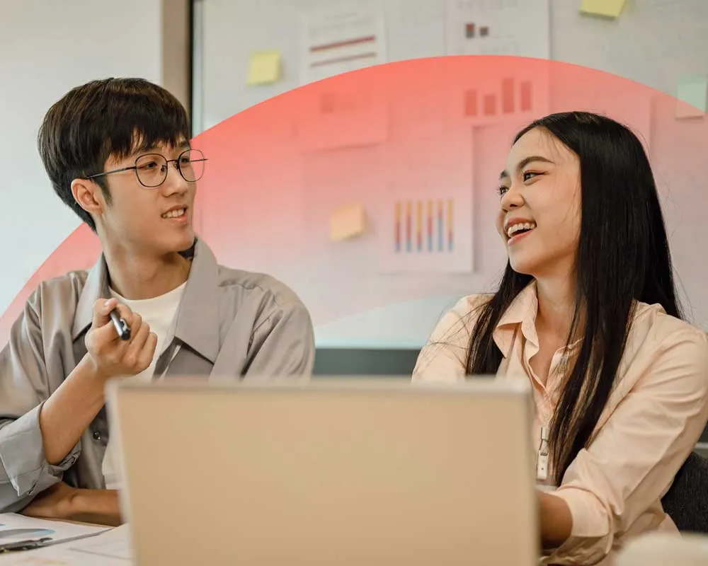 Two colleagues collaborating in an office, smiling and discussing ideas while using a laptop.