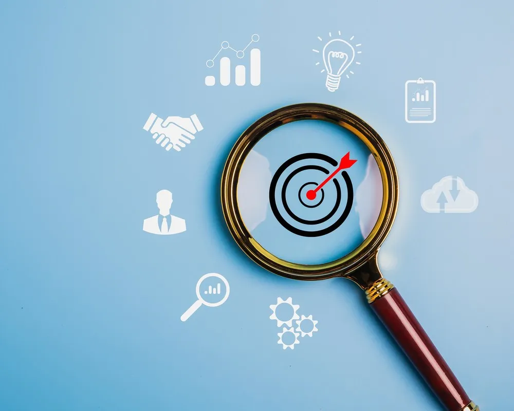 Magnifying glass focused on bullseye target with business icons floating around on blue background