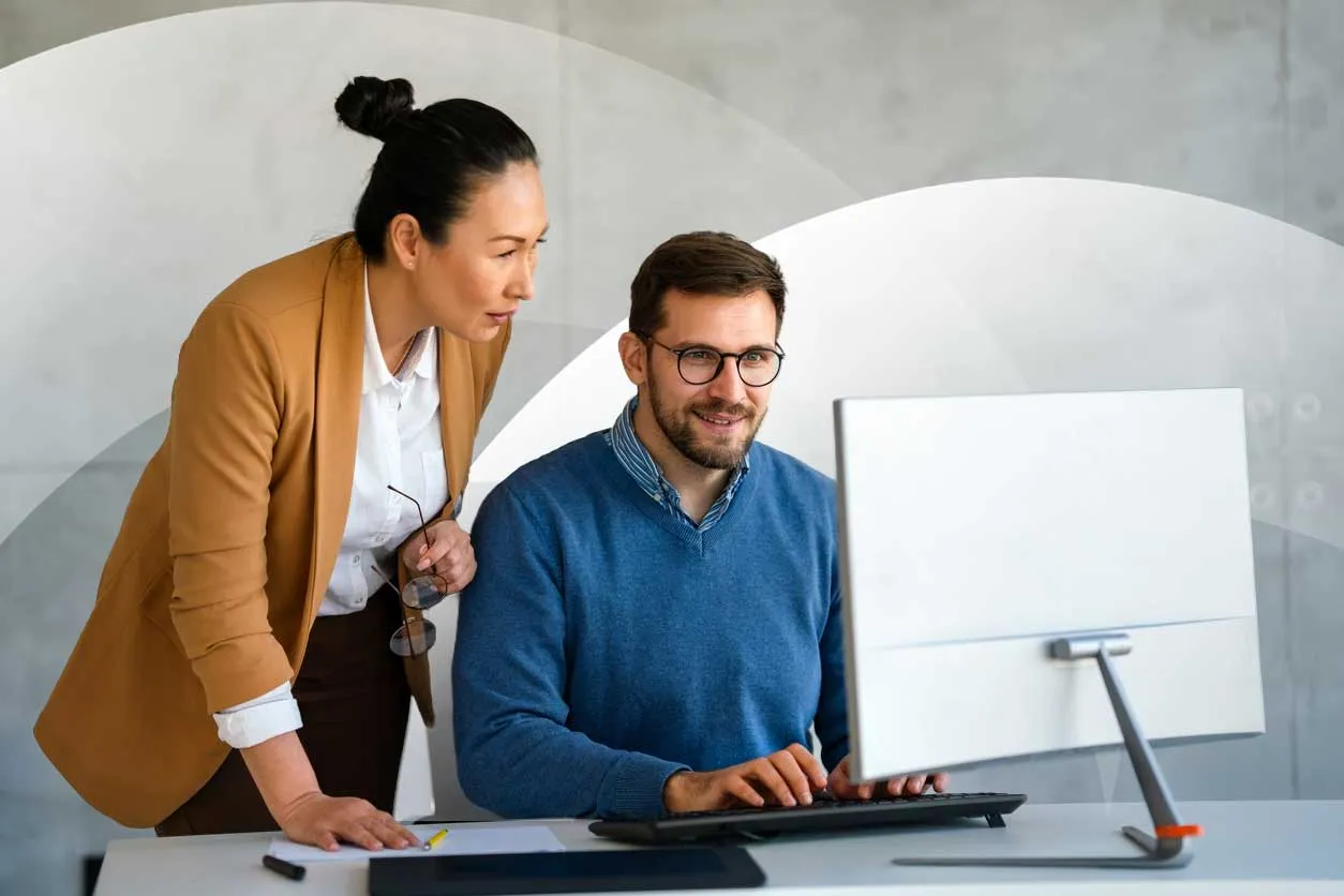 A woman standing beside a seated colleague, both focused on a computer screen while working together