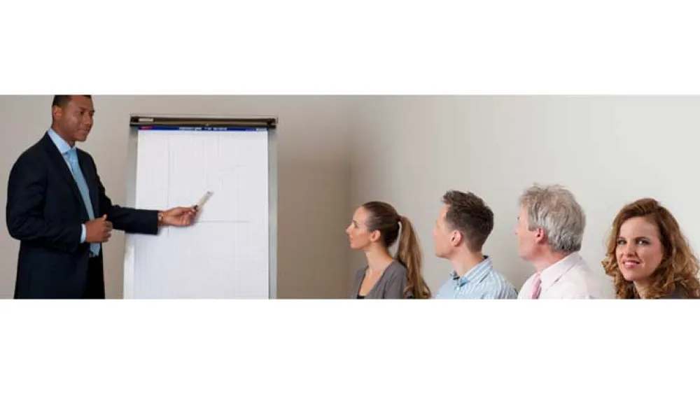 Man pointing at a whiteboard with a captive audience. One lady is looking towards the camera, not paying attention.