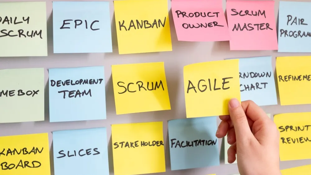 Colorful sticky notes on wall with business terms like 'Epic', 'Agile', and 'Team', hand placing note
