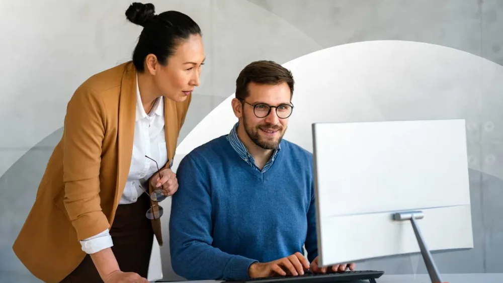 A woman standing beside a seated colleague, both focused on a computer screen while working together