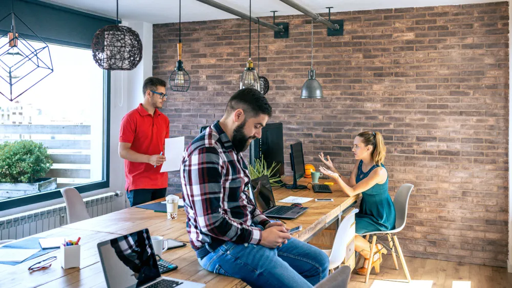 Three people working and chatting in a bright modern office with brick walls