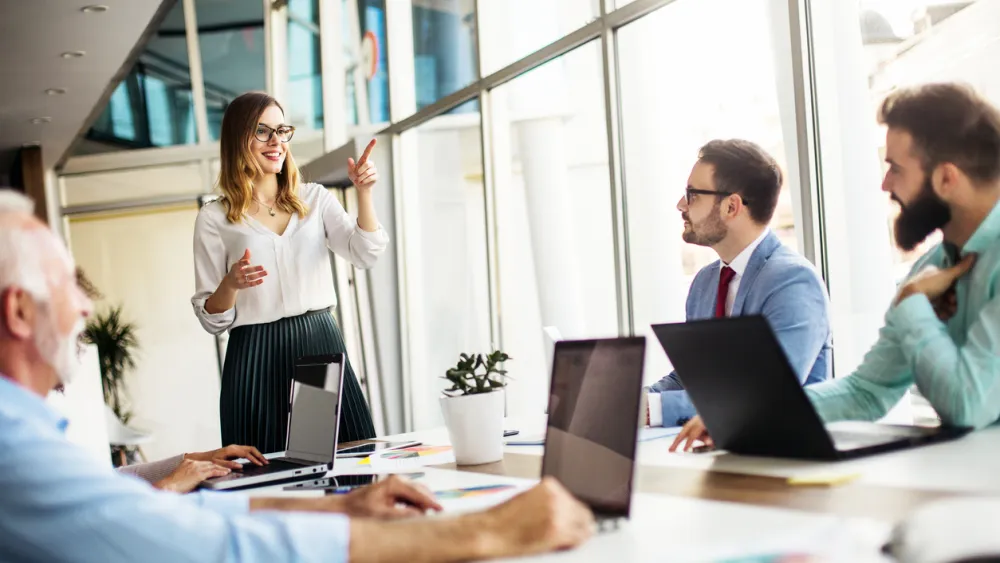 Men sat at a meeting room table with open laptops focus their attention on a woman who stands as she explains coaching as a manager.