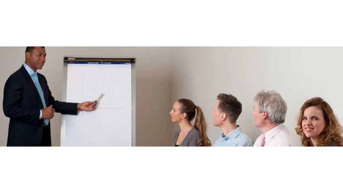 Man pointing at a whiteboard with a captive audience. One lady is looking towards the camera, not paying attention.